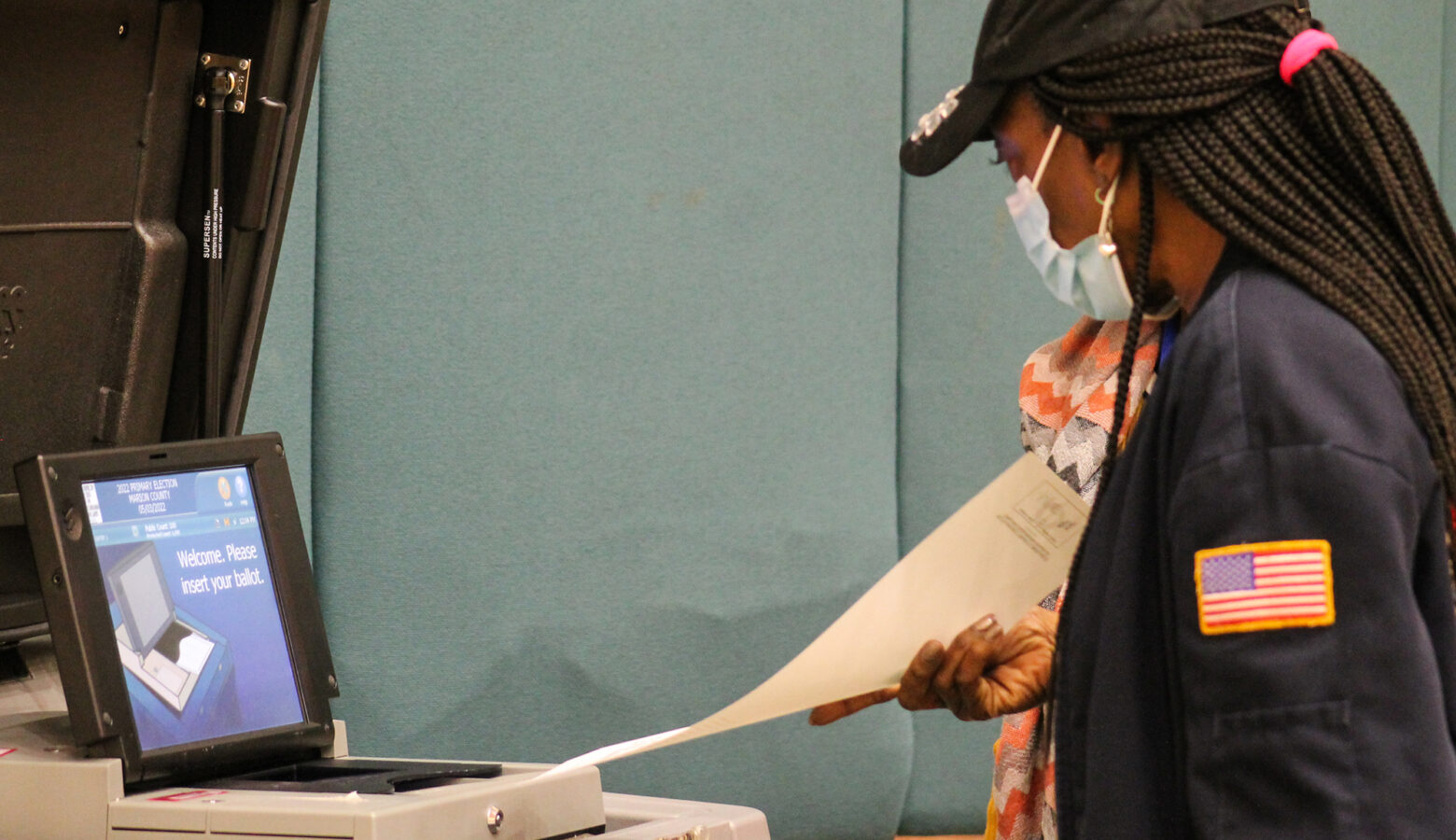 A person wearing a mask feeds their ballot into a return box.
