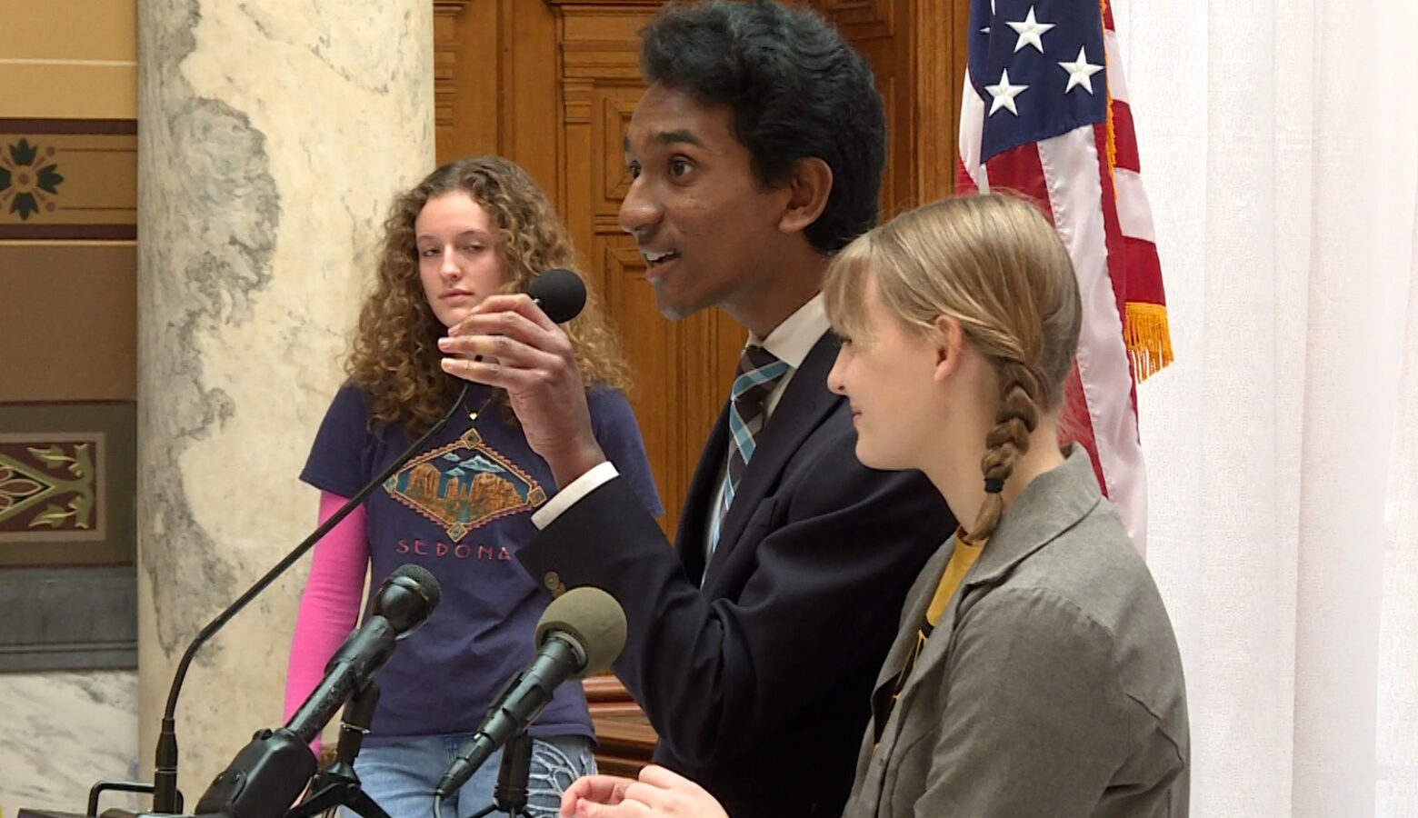 Two youth activists stand near a podium in the Indiana Statehouse. Behind them, large stone columns in the Statehouse atrium are visible, along with an American flag. Another youth activist can be seen in the background, behind the speakers.