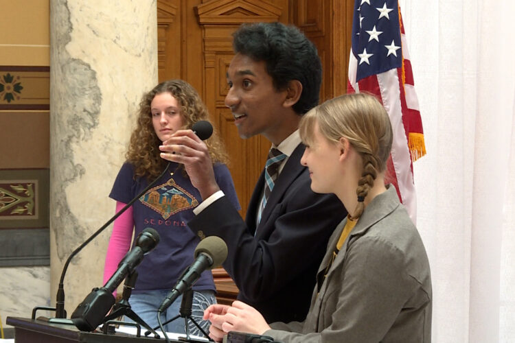 Two youth activists stand near a podium in the Indiana Statehouse. Behind them, large stone columns in the Statehouse atrium are visible, along with an American flag. Another youth activist can be seen in the background, behind the speakers.