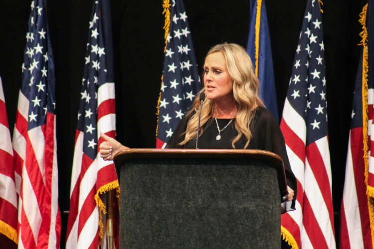 Jennifer McCormick speaks at a podium at the Indiana Democratic State Convention. Behind her is a black curtain with a line of American flags. McCormick is a White woman with blonde hair. She is wearing a black top, with a necklace and large pendant.