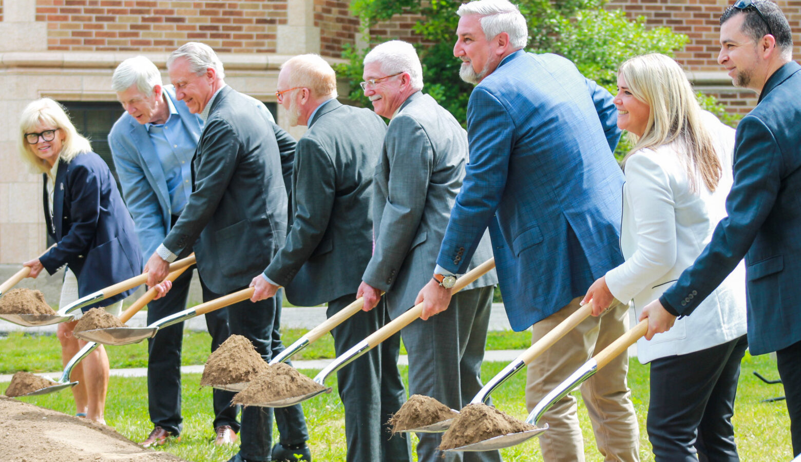 A row of people in suits hold shovels loaded with dirt over a mound.