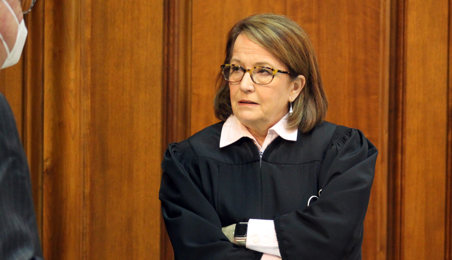 Loretta Rush stands with her arms folded while speaking to someone on the Indiana House floor. Rush is a White woman with brunette hair. She is wearing glasses and black judicial robes over a light-colored shirt.