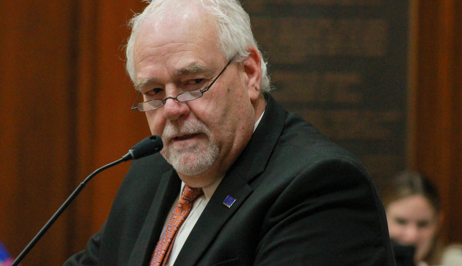 Matt Lehman speaks into a microphone on the floor of the Indiana House. Lehman is a White man with white hair. He is wearing glasses and a black suit.