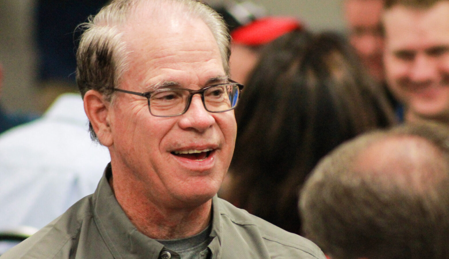 Mike Braun smiles while seated at a breakfast event. Braun is a White man, balding with dark, graying hair. He is wearing glasses and a khaki green shirt.