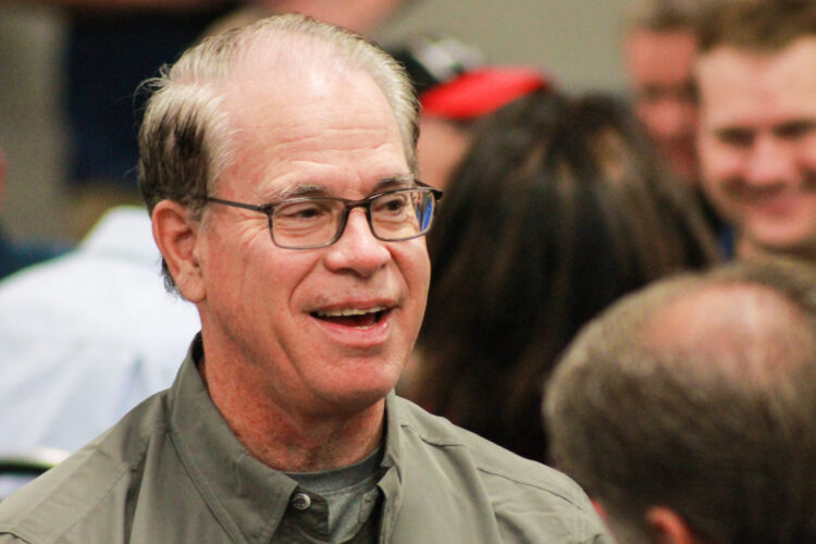 Mike Braun smiles while seated at a breakfast event. Braun is a White man, balding with dark, graying hair. He is wearing glasses and a khaki green shirt.