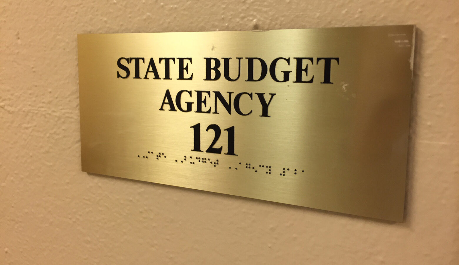 A sign on a wall reads "State Budget Agency" along with the room number. The sign is also written in Braille.