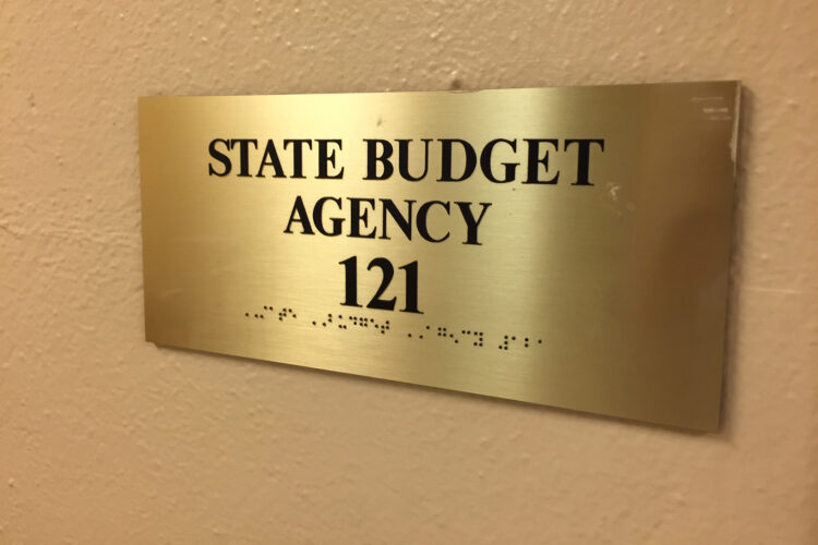 A sign on a wall reads "State Budget Agency" along with the room number. The sign is also written in Braille.