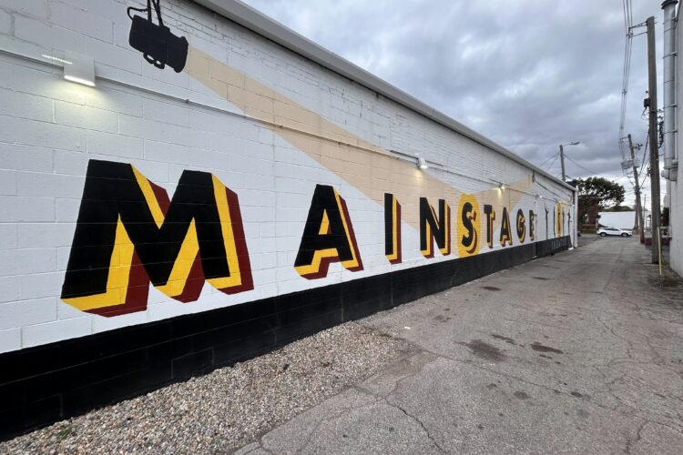 A lettered mural is painted on the outside of a long white building, with the word Mainstage in black and yellow letters.