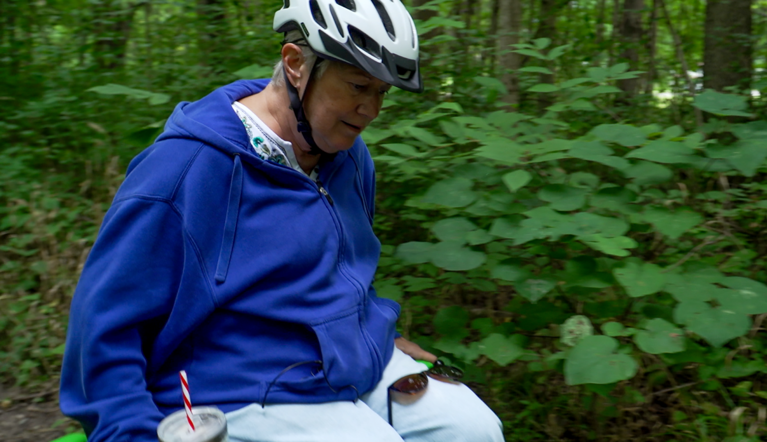 Nancy navigates her trail chair named Peggy in Eagle Creek. She wears a blue zip-up hoodie and light blue pants. She also wears a helmet on the trail with a drink sitting in a cupholder on the chair. Behind her is lush green foliage.