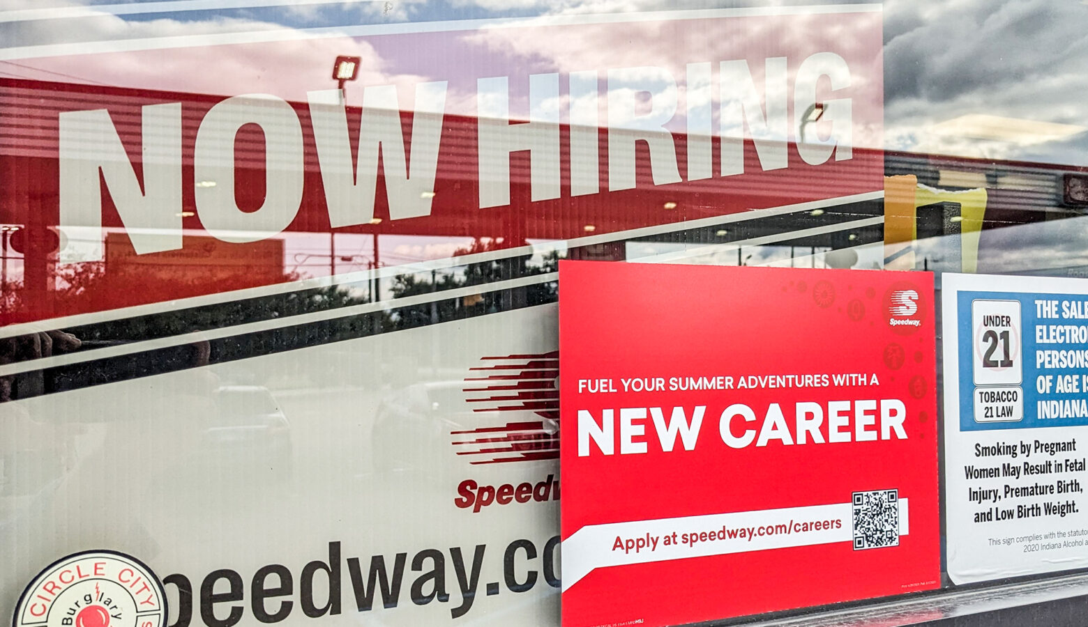 Red sign that reads NOW HIRING in white lettering inside of a window. It's partially covered by a sticker that reads NEW CAREER