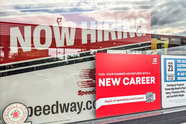 Red sign that reads NOW HIRING in white lettering inside of a window. It's partially covered by a sticker that reads NEW CAREER