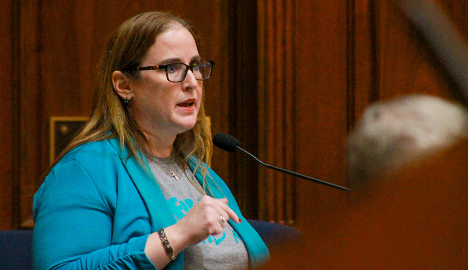 Amber Davis testifies in the Indiana House. She is speaking into a microphone at a lectern. Davis is a White woman with light brown hair. She is wearing glasses and a teal sweater over a gray t-shirt.