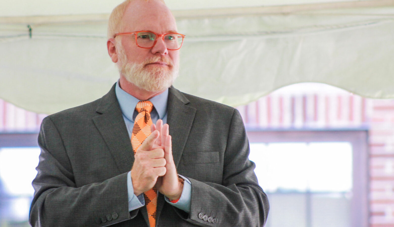 David Geeslin signs while making a speech. Geeslin is a White man with red and white hair and beard. He is wearing glasses and a gray suit, blue shirt and orange checkered tie.