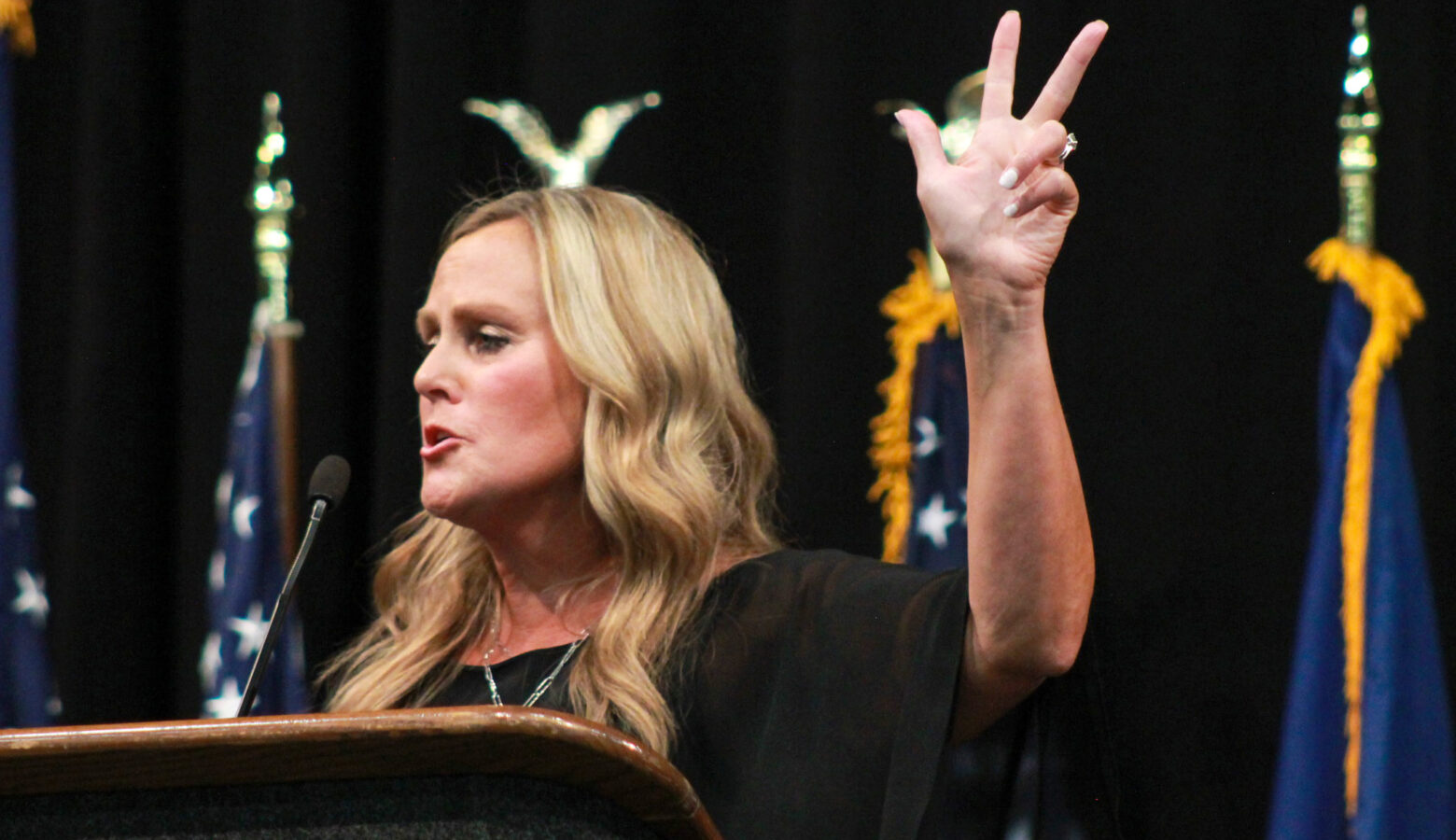 Jennifer McCormick holds her hand in the air, gesturing as she speaks at a lectern. There are a row of flags behind her. McCormick is a White woman with blonde hair. She is wearing a black top.