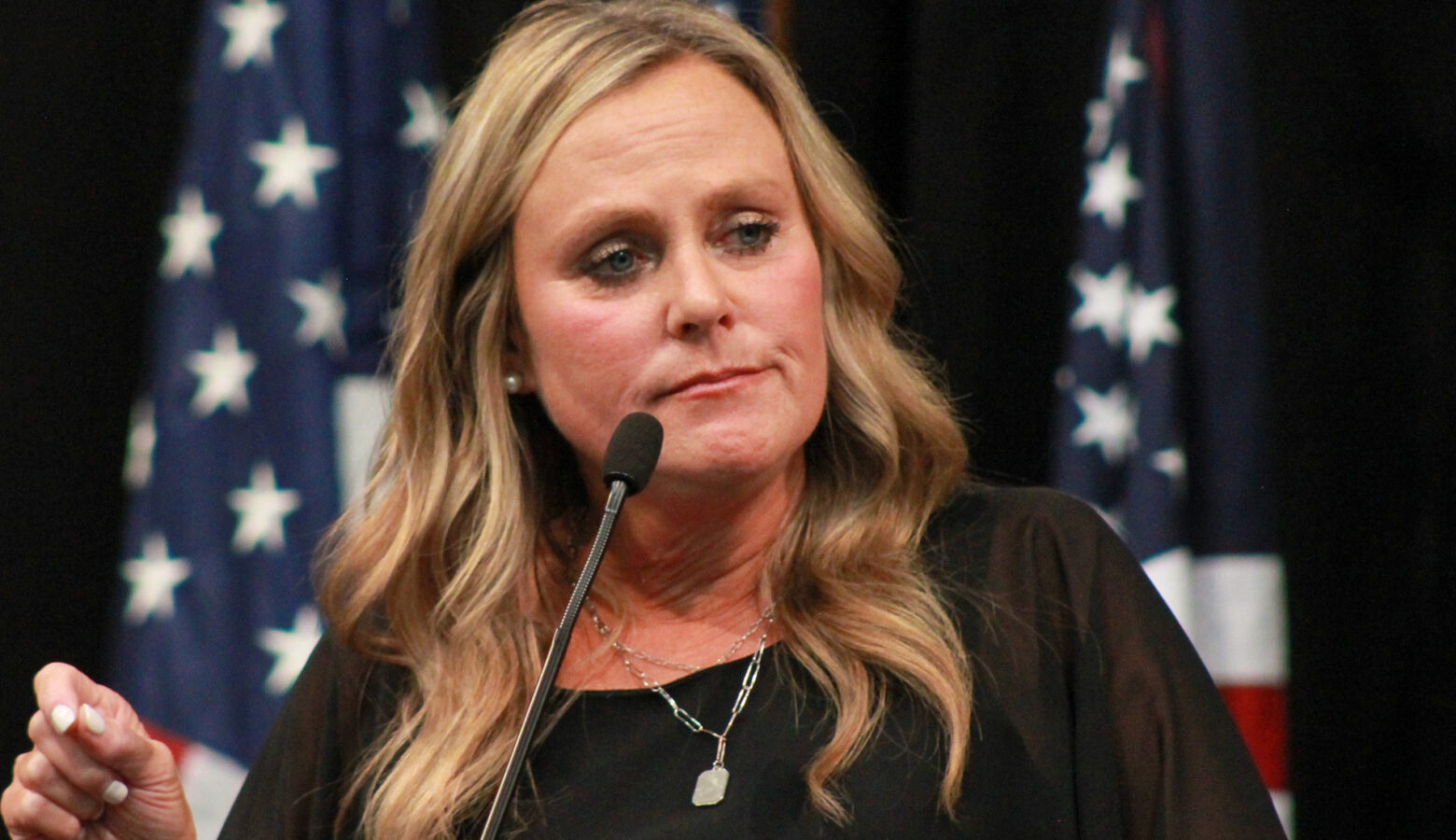 Jennifer McCormick speaks into a microphone on a stage with American flags standing behind her. McCormick is a White woman with blonde hair. She is wearing a black top.