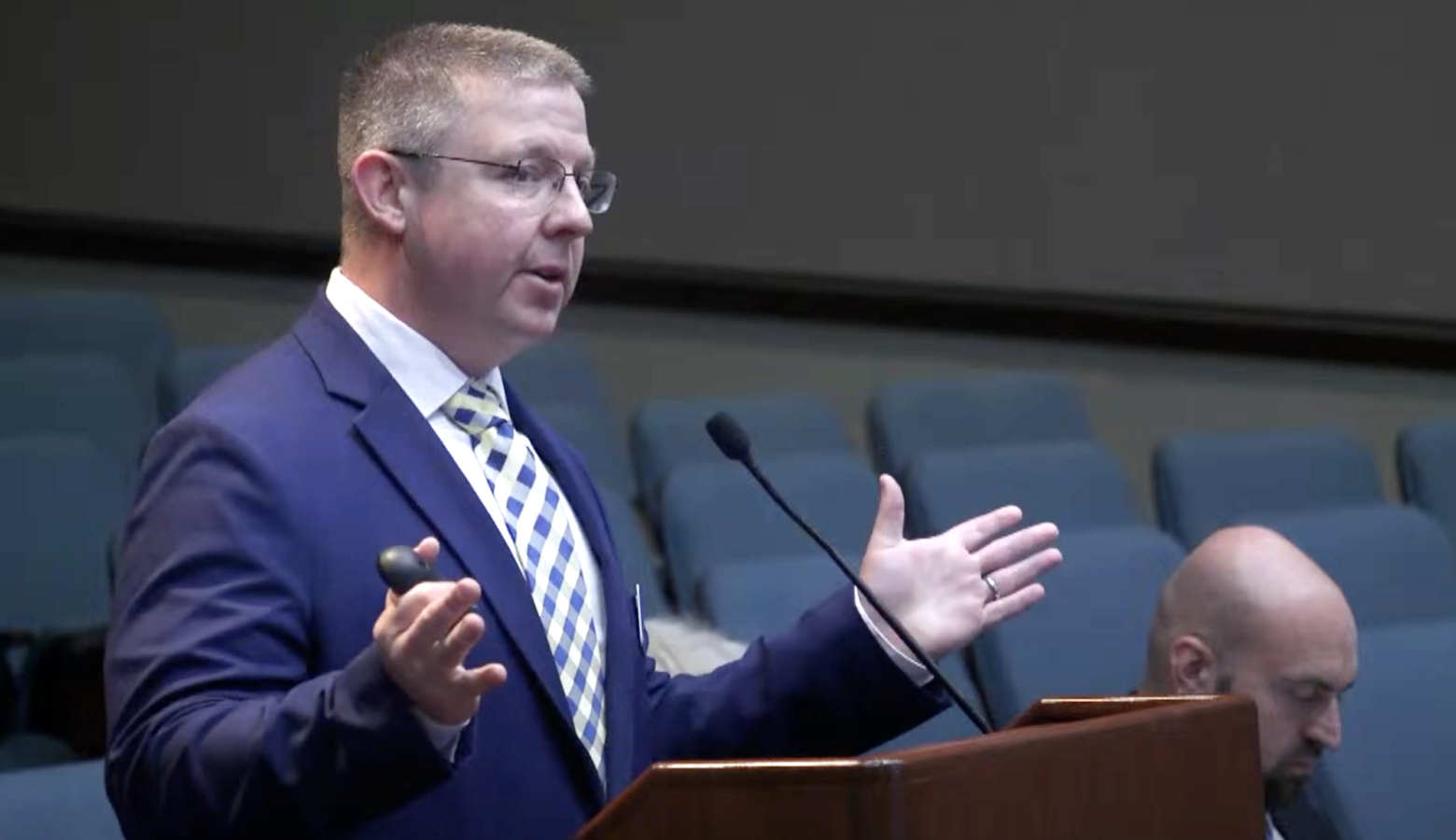 A man in a blue suit with outstretched hands stands in front of a podium and speaks into a microphone.