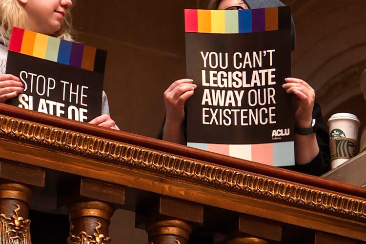 Protesters at the Statehouse hold up signs that read "You Can't Legislative Away Our Existence" with the ACLU of Indiana logo.