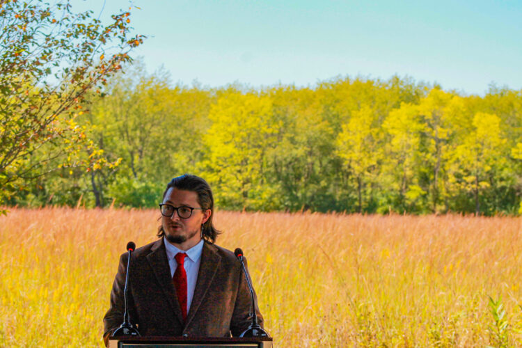 Logan York stands at a lectern in front of prairie grass and, further in the background, a forest. York is a man with long, dark hair and a beard. He is wearing glasses and a brown sport coat over a shirt and tie.