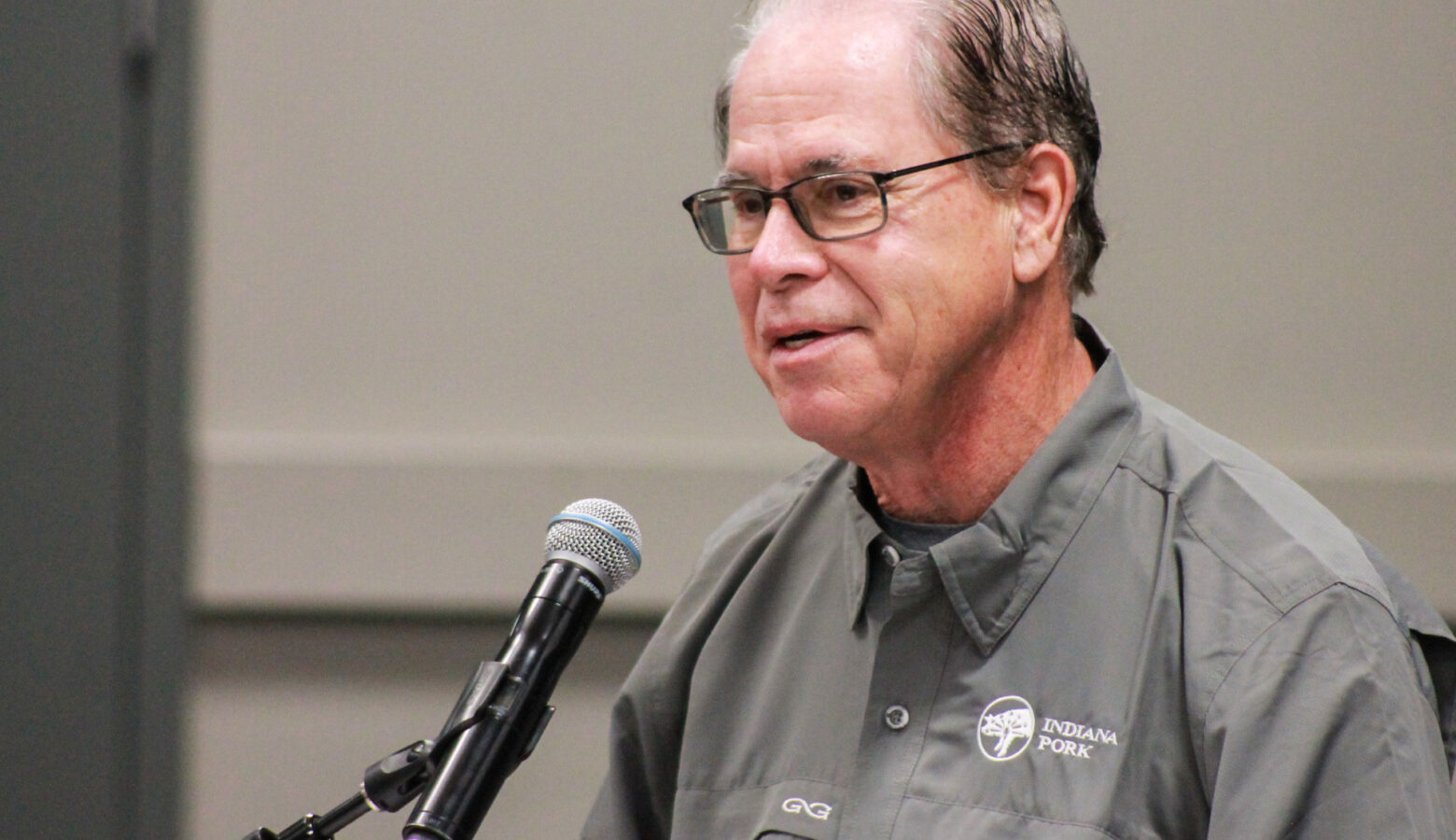 Mike Braun speaks into a microphone. Braun is a White man, balding with dark gray hair. He is wearing glasses and a button-down shirt with the Indiana Pork Producers logo.