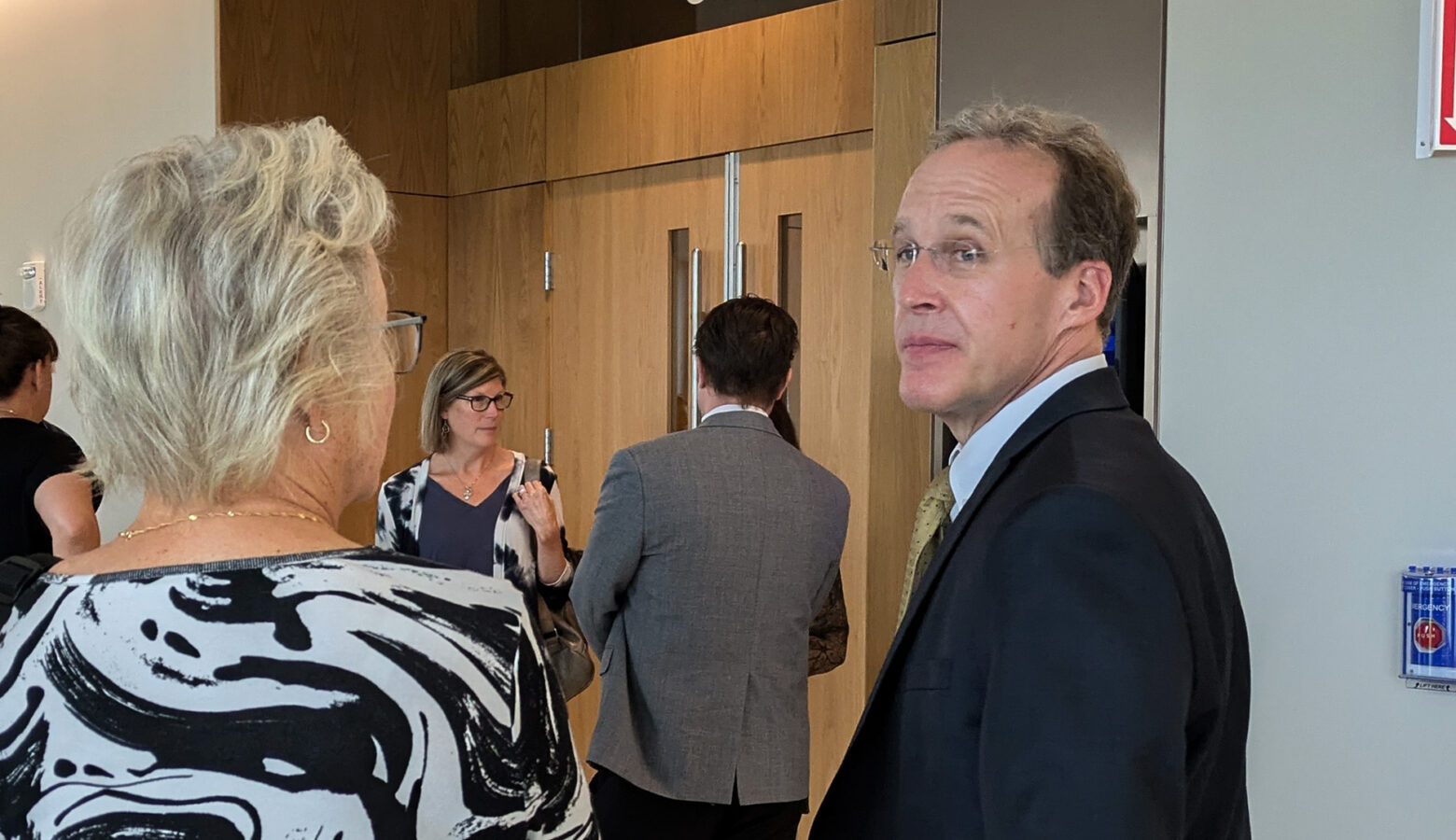 Patrick Gillen glances over his shoulder outside of the courtroom. He is speaking to a woman with light hair and a black and white shirt.