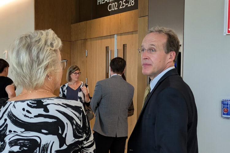 Patrick Gillen glances over his shoulder outside of the courtroom. He is speaking to a woman with light hair and a black and white shirt.