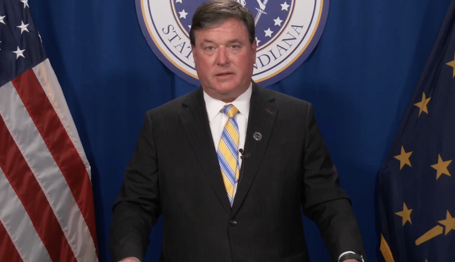 A screenshot of a Facebook Live shows Todd Rokita standing at a lectern in front of the American and Indiana flags, with the seal of the office of the attorney general behind him. Rokita is a White man with dark, graying hair. He is wearing a suit and tie.