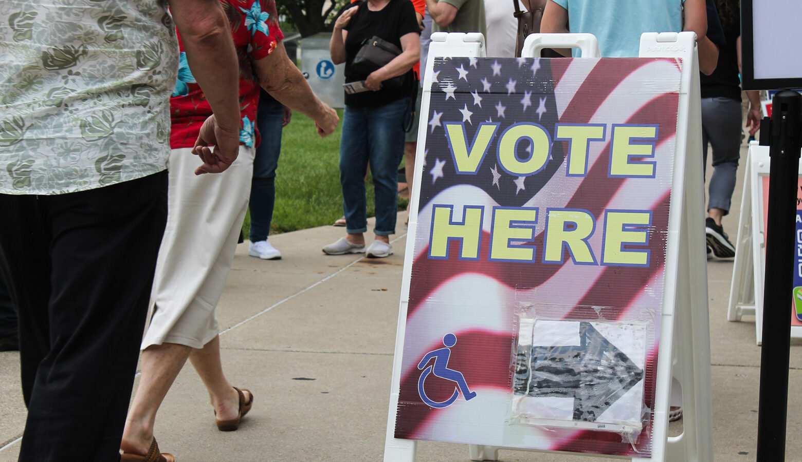 A sidewalk sign reads "Vote Here" with an arrow pointing to the right and a handicap accessibility logo. There are people walking by the sign.