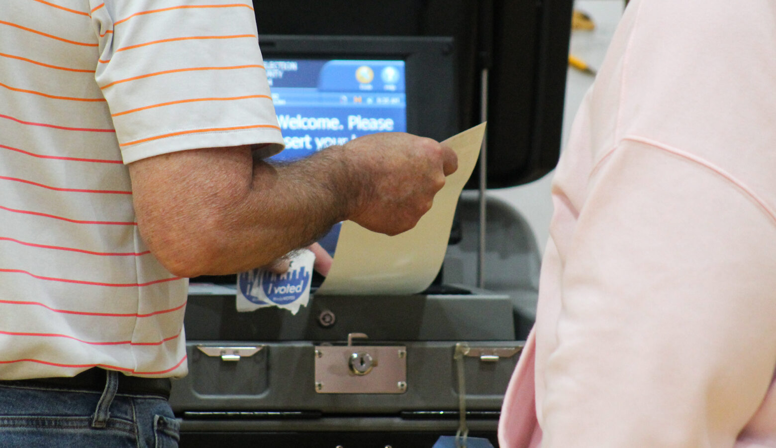 A person feeds a paper ballot into a machine as an election worker is available to assist.