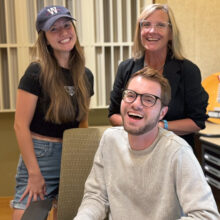 Three people stand together, with one sitting at a desk.