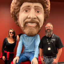 A woman and a man pose next to a large statue of Bob Ross at the IPR Studios.