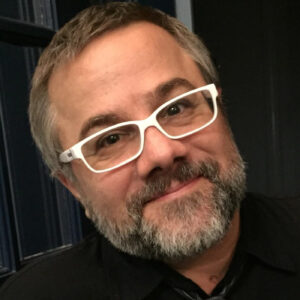 A man with a salt-and-pepper beard and rectangular white glasses smiles at the camera.