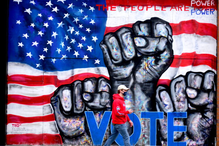 A person in a red hoodie walk past a wall mural. It depicts three raised fists in front of the American Flag with the word "vote."