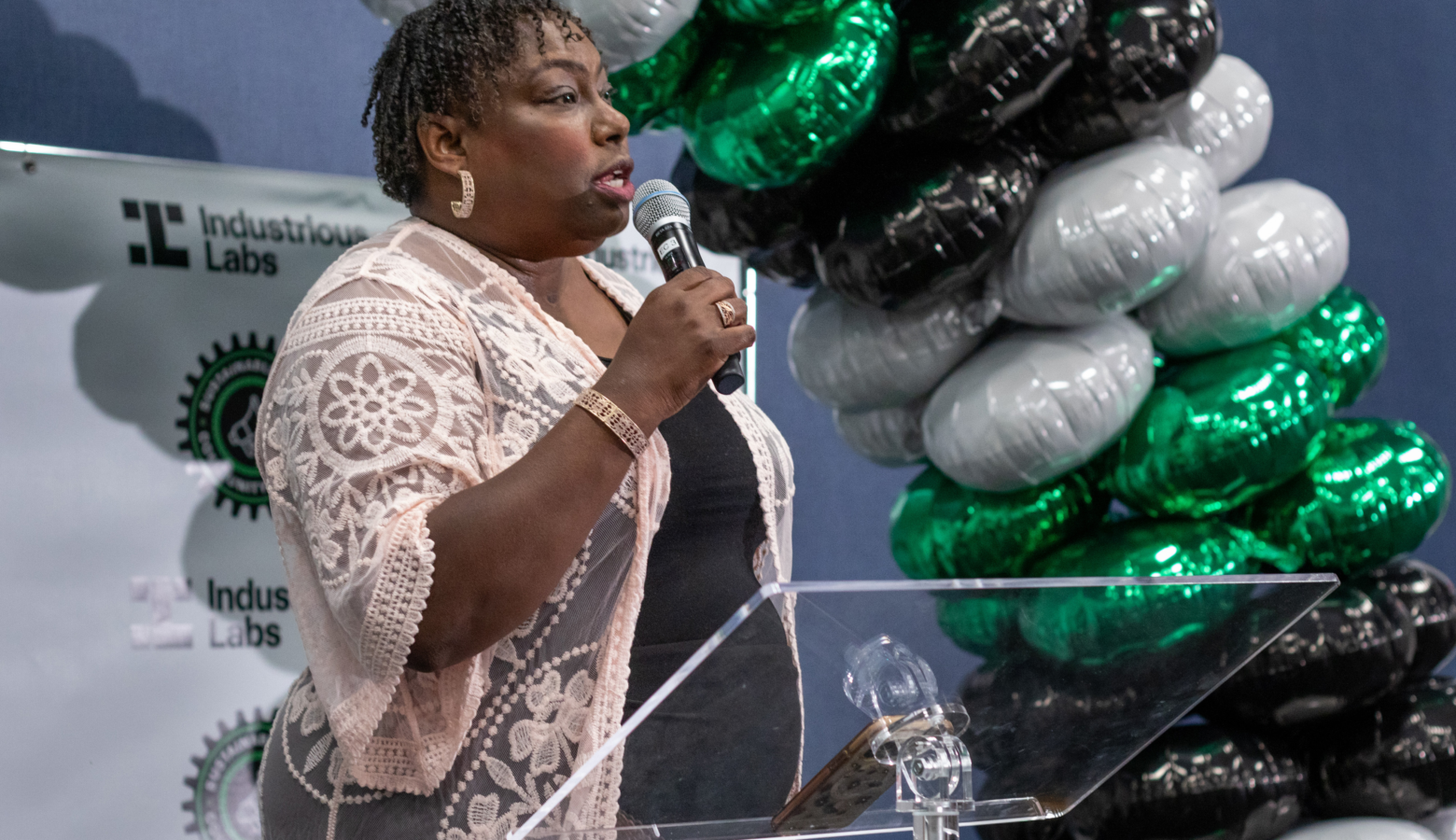 Beverly Lewis holds a microphone at an event. She is a Black woman with short hair, and stands in front of a balloon arch.