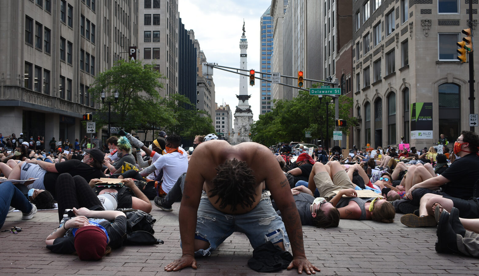 Several dozen protesters lay on the brick roads in downtown Indianapolis as part of a "die in." In the foreground, the main subject of the image is a Black protester, on their hands and knees, shirtless, with their head hung. Directly behind them is Indianapolis's Monument Circle.