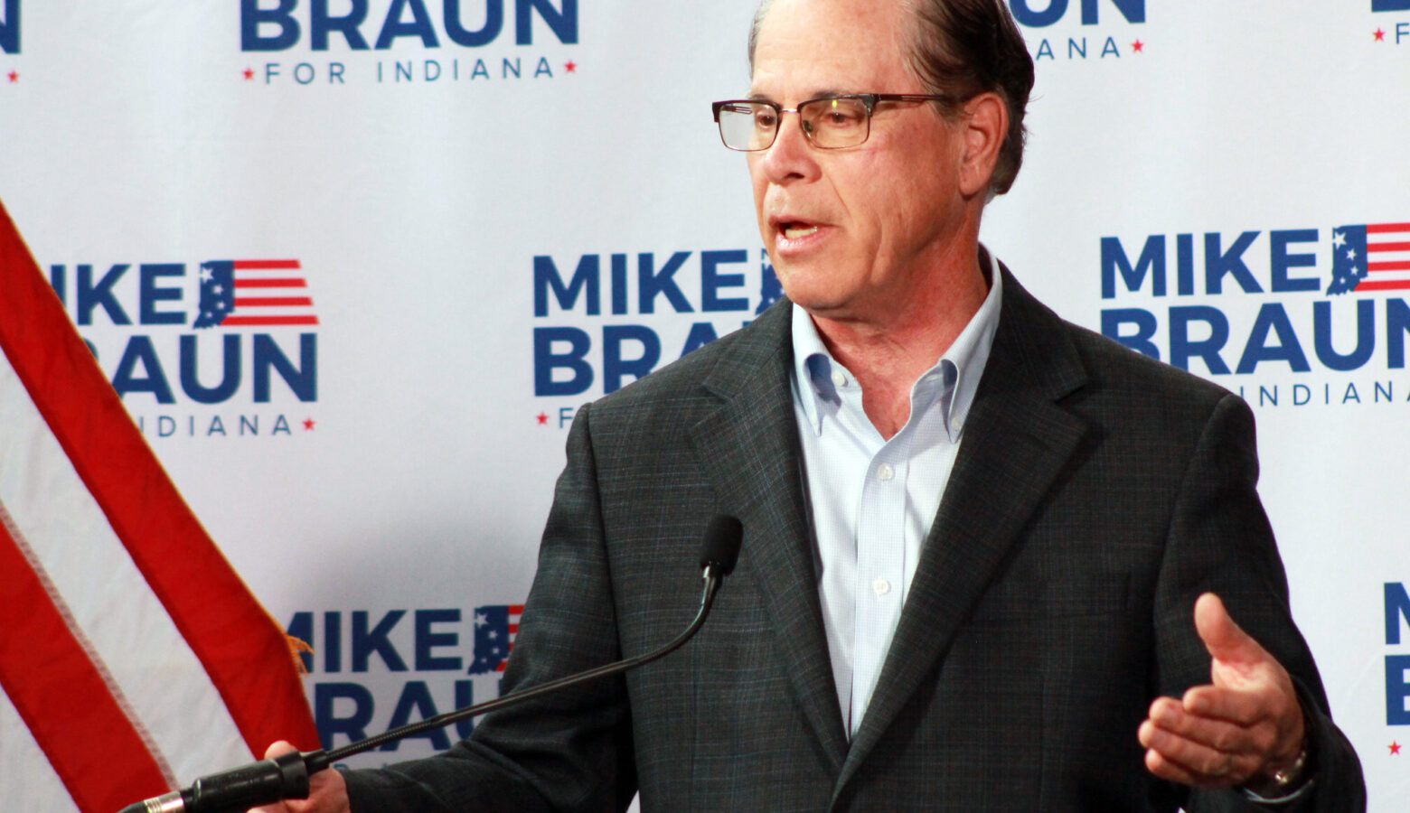 Mike Braun speaks at a lectern. Behind him is a backdrop with his campaign logo dotted across it. Braun is a White man, balding with dark gray hair. He is wearing a dark suit coat and a light-colored shirt.