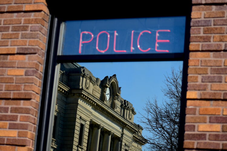 A neon sign reading "Police" hangs in a window. A county courthouse is seen in the window's reflection.