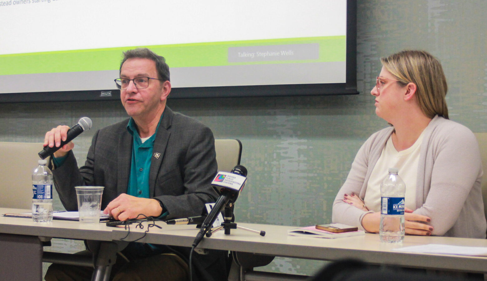 Larry DeBoer is a White man with dark hair. He is wearing glasses and a gray blazer over a green shirt. Neva Butkus is a White woman with blonde hair. She is wearing glasses and a cream sweater over a white top.