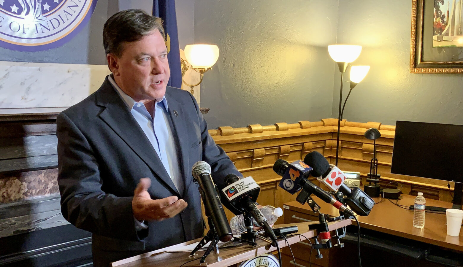 Todd Rokita stands in front of a bank of microphones. Rokita is a White man with dark, graying hair. He is wearing a dark-colored sport coat over a light blue dress shirt.