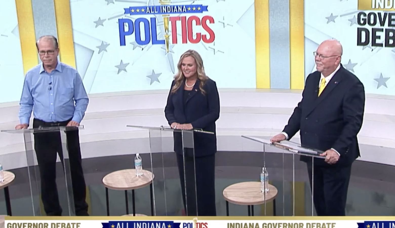 A screenshot of the WISH-TV gubernatorial debate. Mike Braun is a White man with dark, graying hair. He is wearing glasses and a blue shirt with black pants. Jennifer McCormick is a White woman with blonde hair. She is wearing a black pantsuit over a black top. Donald Rainwater is a White man, bad with a white goatee. He is wearing glasses and a black suit with a white shirt and yellow tie.