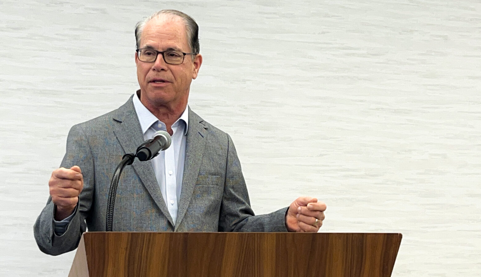 Mike Braun speaks into a microphone at a lectern. Braun is a White man, balding, with gray hair. He is wearing a gray blazer and a light colored shirt.