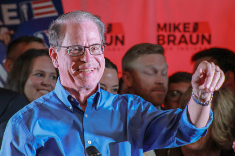 Mike Braun stands on a stage, gesturing while supporters stand behind him. Braun is a White man, balding with gray hair. He is wearing glasses and a blue shirt.